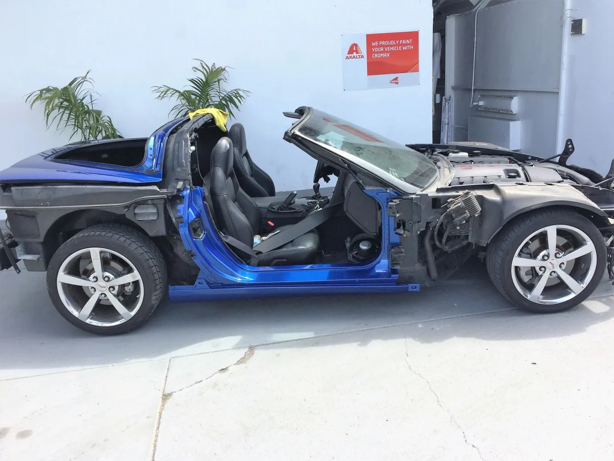 A blue Chevrolet Corvette stripped down to its bare frame and seats inside the Elite Auto Collision bay, with the doors, roof, front clip and body panels all removed and the car sitting on its wheels against a white shop wall