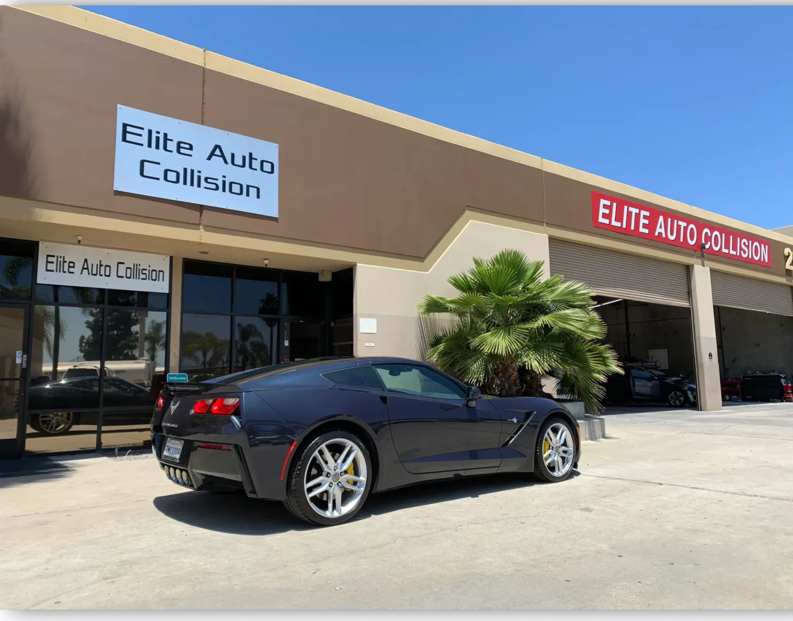 A dark blue Corvette Stingray parked outside the Elite Auto Collision shop on West Bradley Avenue in El Cajon, with open repair bays and the red Elite Auto Collision sign above them